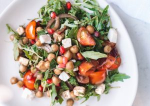 Chickpea, avocado, rocket, tomato, pomegranate, feta and mint salad with balsamic dressing on white plate